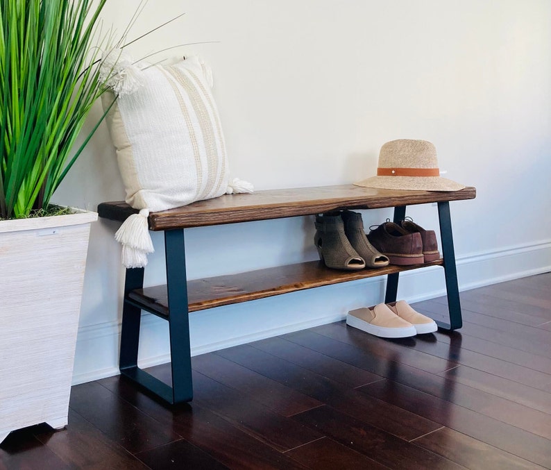 May include: A wooden bench with black metal legs and a shelf. The bench has a white throw pillow and a straw hat on top. There are shoes on the shelf.