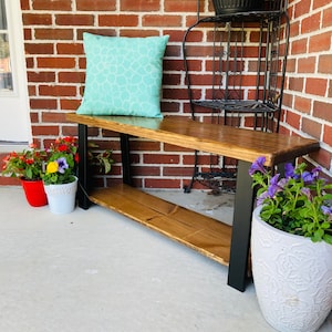 May include: A wooden bench with black metal legs and a teal cushion. The bench is sitting on a concrete patio in front of a brick wall. There are flowers in pots on either side of the bench.