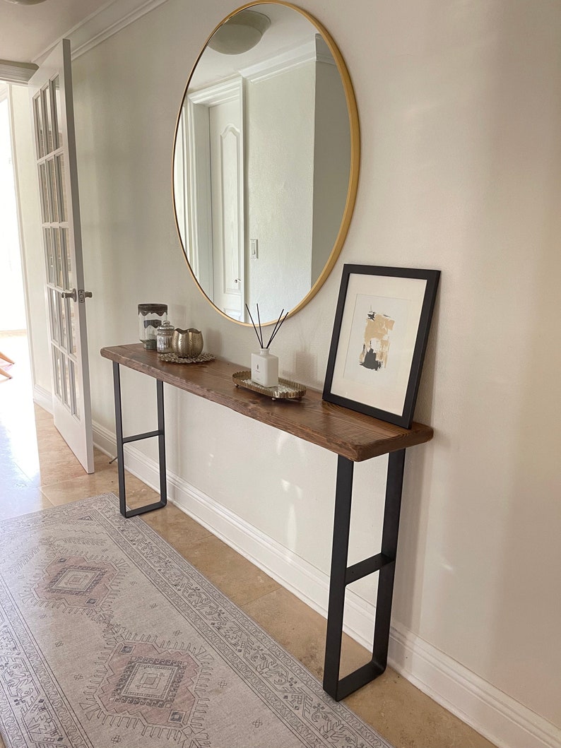 May include: A wooden console table with black metal legs. The table has a live edge top and is decorated with candles, a vase, and a framed print. The table is in front of a large round mirror with a gold frame.