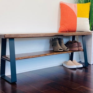 May include: A wooden bench with a black metal frame and a shelf. The bench has a natural wood finish and the shelf is stained a darker brown. There are shoes on the shelf and on the floor.