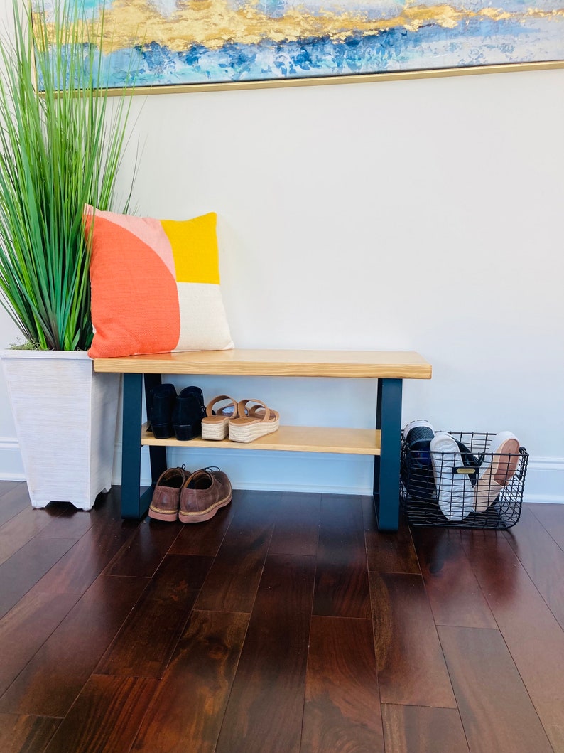 May include: A wooden bench with a black metal frame and a shelf for shoes. The bench has a light wood finish and is sitting on a dark wood floor. There is a pillow on the bench with a geometric pattern in shades of orange, yellow, and white. A black wire basket with shoes is on the floor next to the bench.