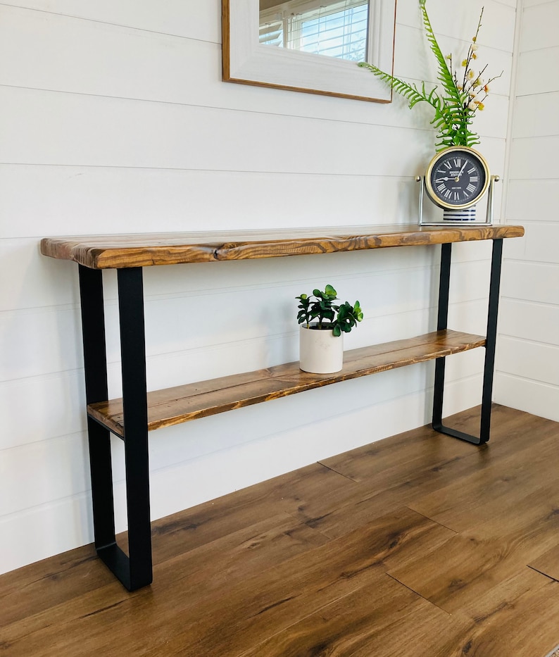 May include: A rustic wooden console table with a black metal frame and two shelves. The table has a natural wood finish and a live edge design. A small potted plant sits on the top shelf.