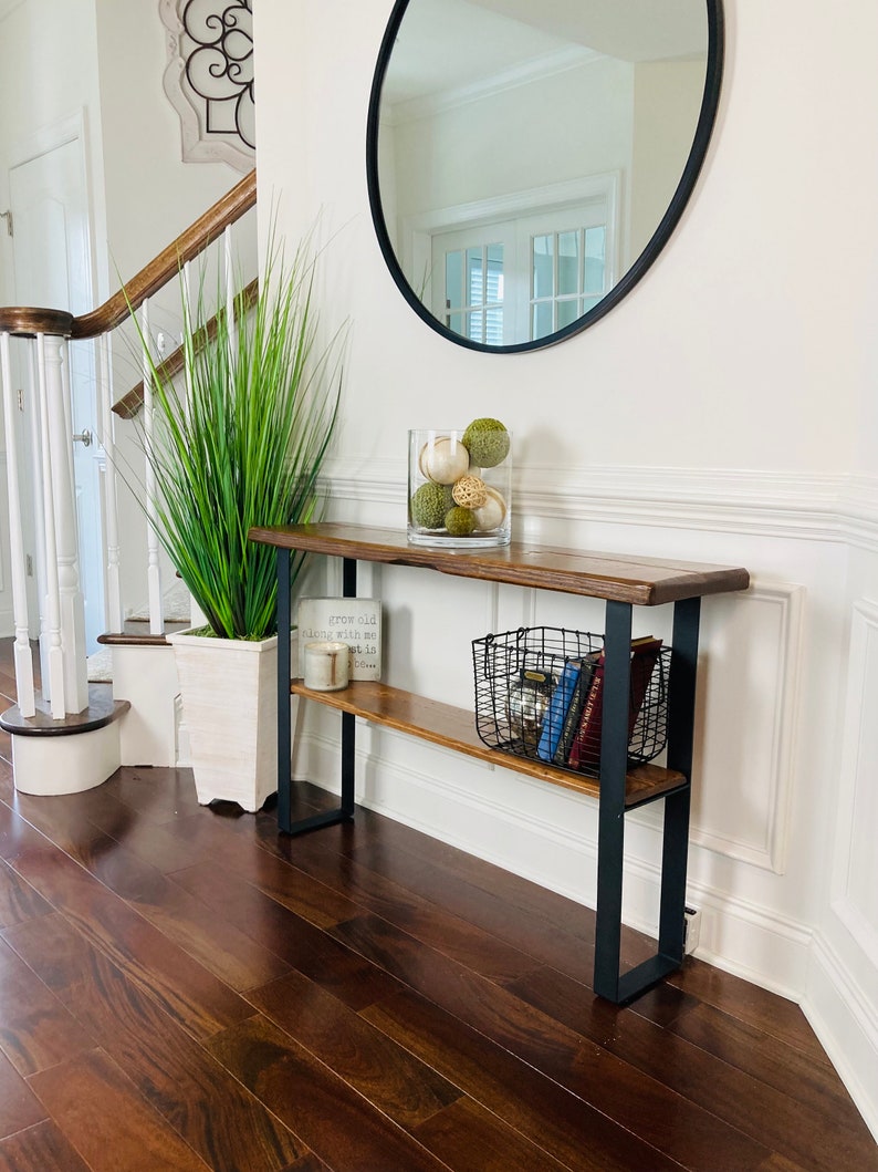 May include: A dark wood console table with a black metal frame and a shelf. The table has a vase with green and white decorative balls on top and a wire basket with books on the shelf. The table is in a hallway with a white wall and a wooden staircase in the background.
