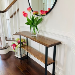 May include: A wooden console table with a black metal frame and two shelves. The table is decorated with a vase of pink tulips and a small arrangement of pink flowers.