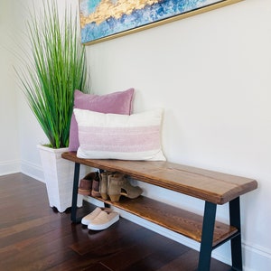 May include: A wooden bench with a black metal frame and a shelf underneath. The bench has two pillows on top and shoes are on the shelf.