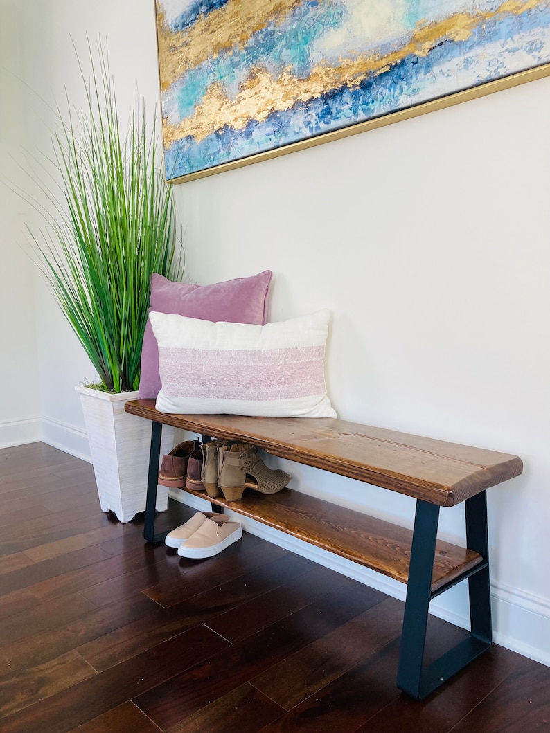 May include: A wooden bench with a black metal frame and a shelf underneath. The bench has two pillows on top and shoes are on the shelf.