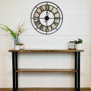 May include: A rustic wooden console table with a black metal frame and a shelf. The table has a distressed wood finish and is decorated with a large, round clock with Roman numerals.