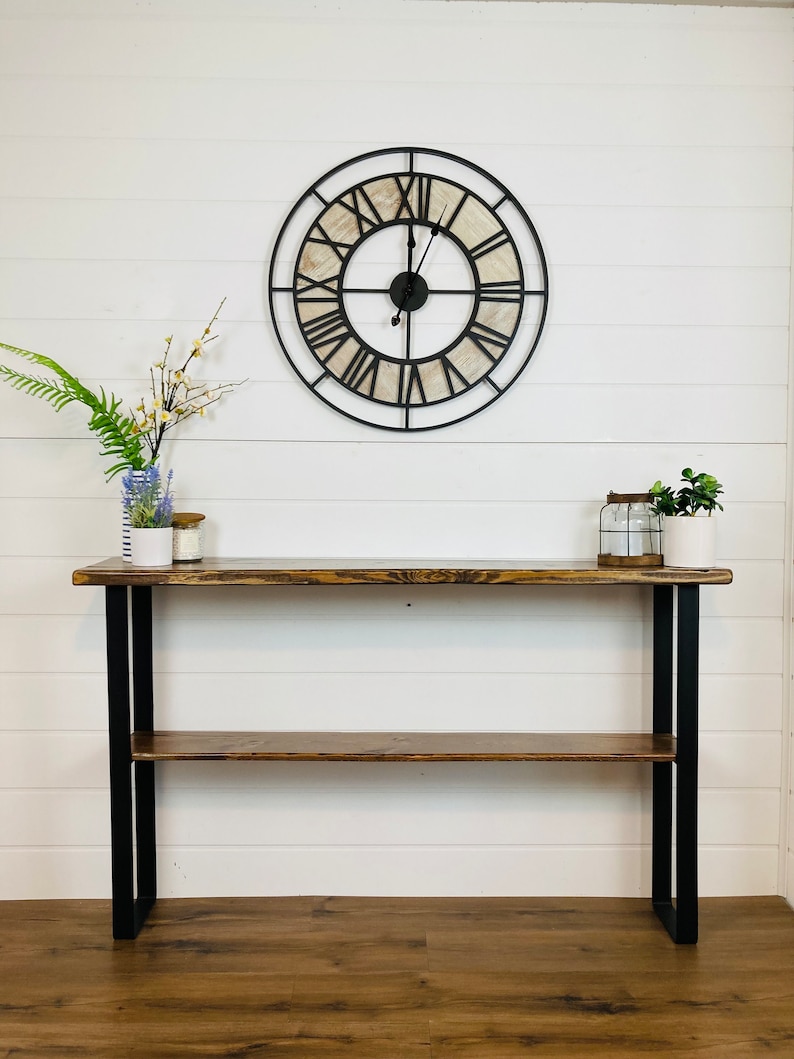 May include: A rustic wooden console table with a black metal frame and a shelf. The table has a distressed wood finish and is decorated with a large, round clock with Roman numerals.