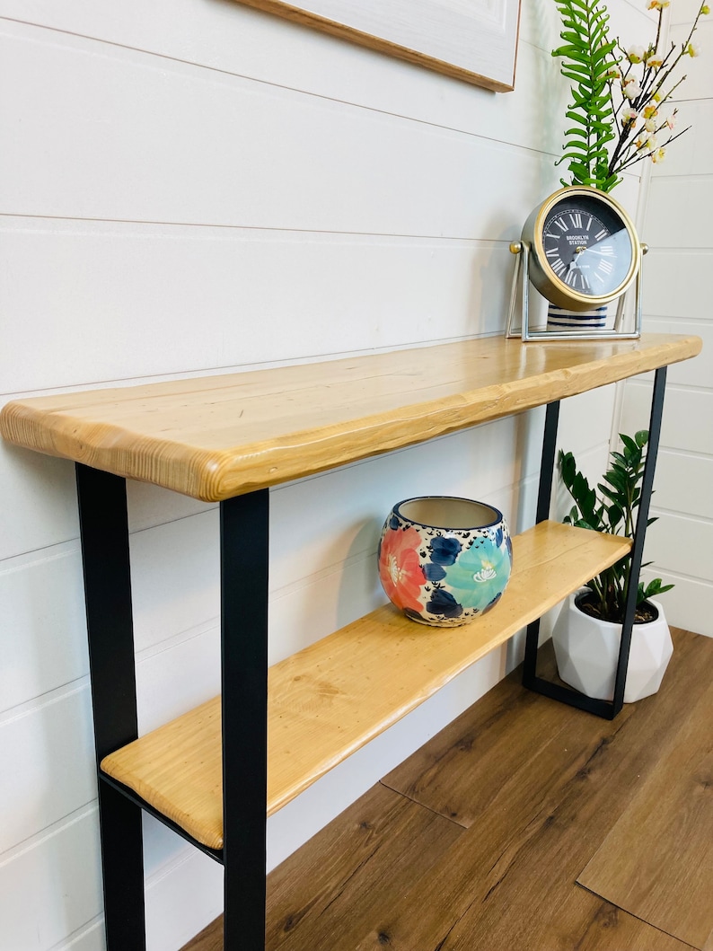 May include: A wooden console table with a live edge top and black metal legs. The table has a shelf and is decorated with a floral patterned ceramic pot and a clock.