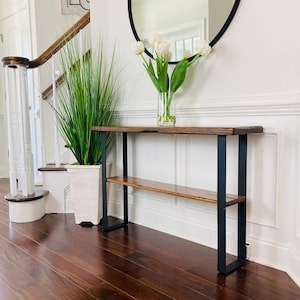 May include: A wooden console table with a black metal frame and a lower shelf. The table is placed in a hallway with a white wall and a round mirror above it. A vase of white tulips is on the table.