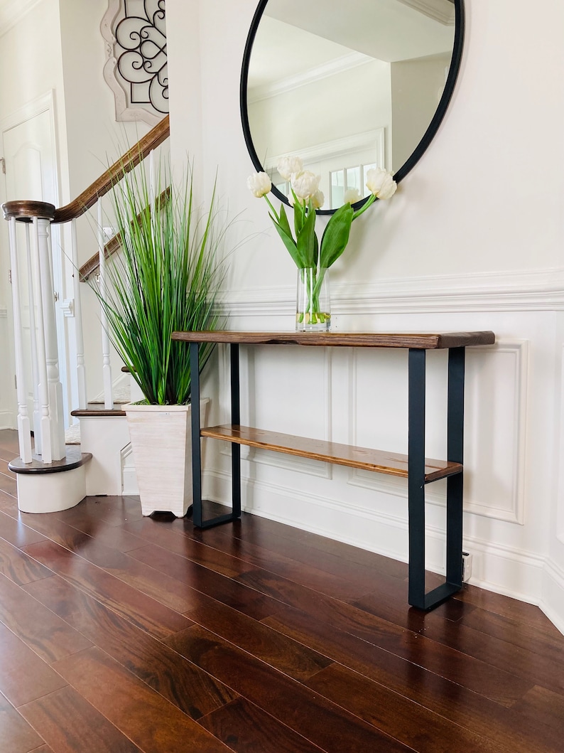 May include: A wooden console table with a black metal frame and a lower shelf. The table is placed in a hallway with a white wall and a round mirror above it. A vase of white tulips is on the table.