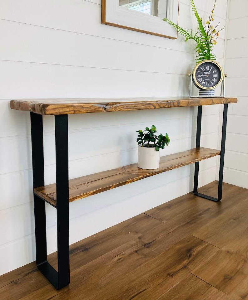 May include: A rustic wooden console table with a black metal frame and a lower shelf. The table has a natural wood finish and a distressed look. It is decorated with a potted plant and a clock.