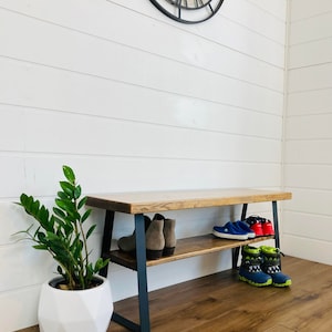 May include: A wooden bench with a black metal frame and two shelves. The bench has a dark brown stain and is sitting on a light brown wood floor. There are shoes on the bottom shelf and a potted plant on the floor next to the bench.