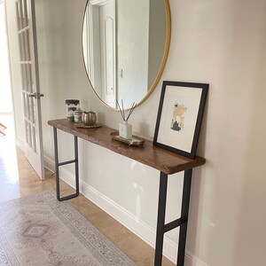 May include: A wooden console table with black metal legs. The table is decorated with candles, a gold bowl, and a diffuser. A round gold mirror hangs on the wall above the table. A framed abstract art print hangs on the wall to the right of the table.