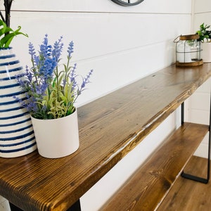 May include: A rustic wooden console table with a dark brown stain and black metal legs. The table has a single shelf and is decorated with two potted plants and a lantern.