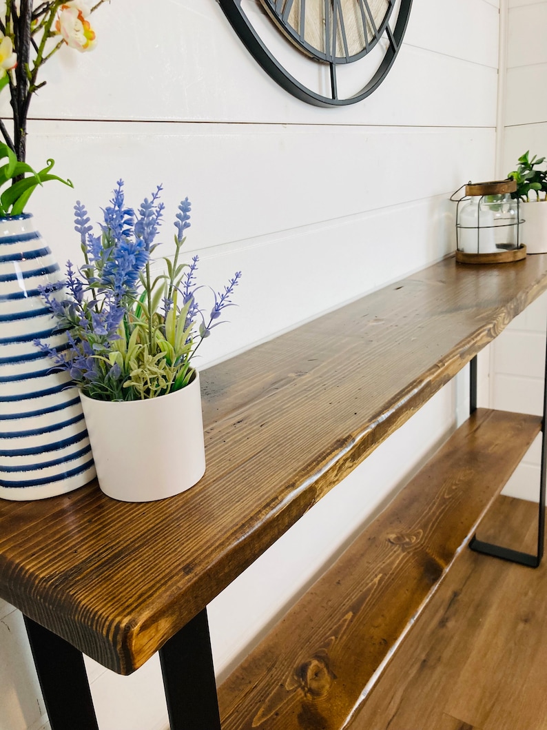 May include: A rustic wooden console table with a dark brown stain and black metal legs. The table has a single shelf and is decorated with two potted plants and a lantern.