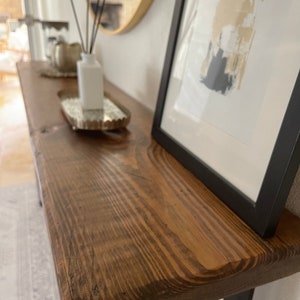 May include: A wooden console table with a natural wood finish and black metal legs. The table is decorated with a silver tray, a white bottle, and a framed abstract print.