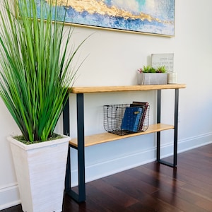 May include: A wooden console table with black metal legs and two shelves. The top shelf has a black wire basket with books inside. The bottom shelf is empty. A white planter with a green plant is on the floor to the left of the table. A framed abstract painting is on the wall behind the table. A small sign with the text "grow old along with me the best is yet to be" is on the top shelf.