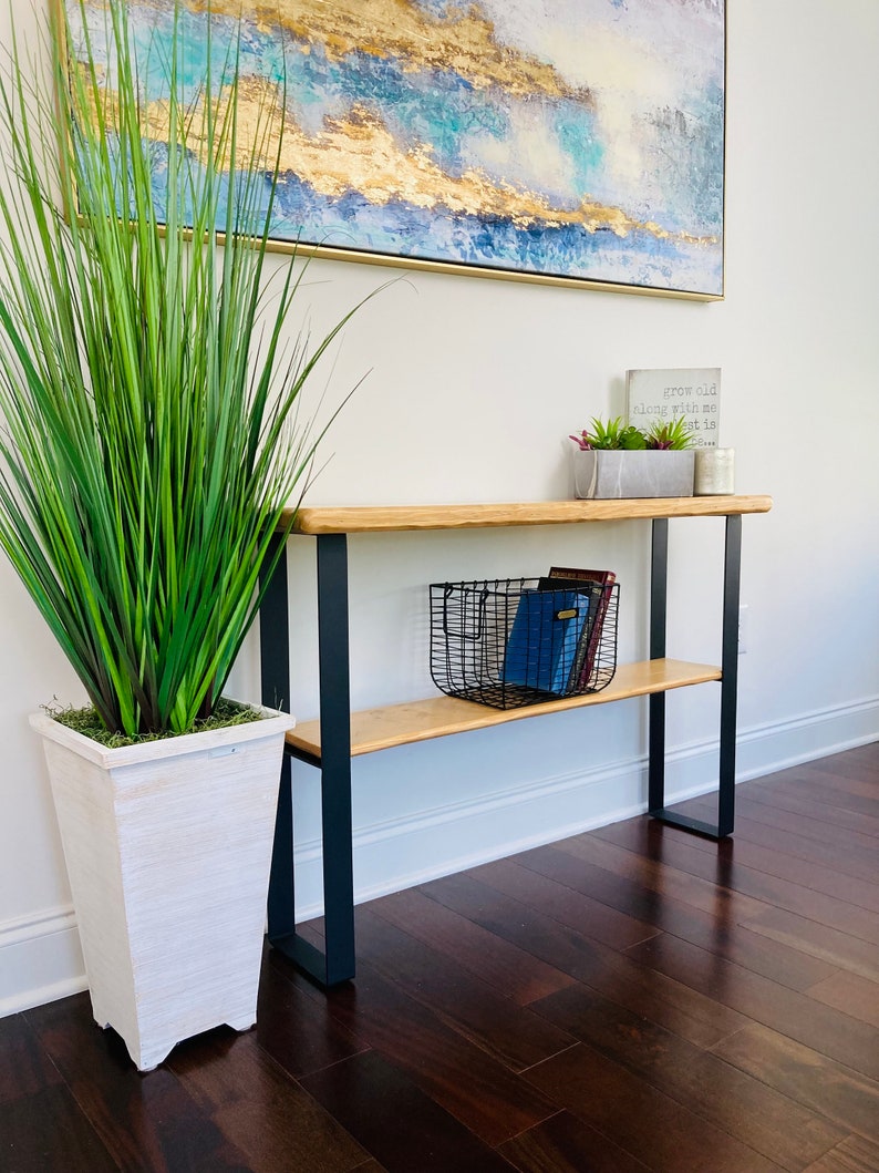 May include: A wooden console table with black metal legs and two shelves. The top shelf has a black wire basket with books inside. The bottom shelf is empty. A white planter with a green plant is on the floor to the left of the table. A framed abstract painting is on the wall behind the table. A small sign with the text "grow old along with me the best is yet to be" is on the top shelf.