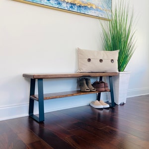 May include: A wooden bench with a black metal frame and a shelf underneath. The bench has a natural wood finish and is sitting on a dark wood floor. There are shoes on the shelf and a pillow on the bench.