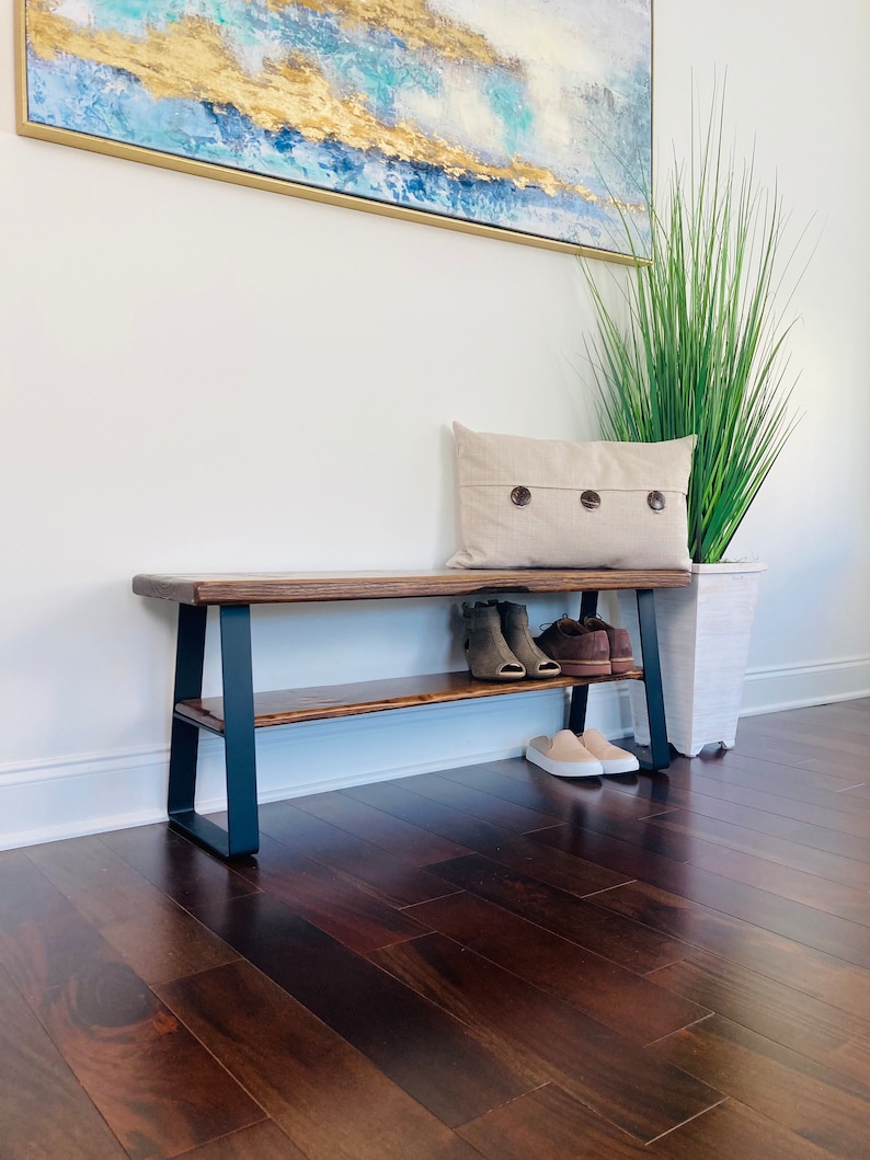 May include: A wooden bench with a black metal frame and a shelf underneath. The bench has a natural wood finish and is sitting on a dark wood floor. There are shoes on the shelf and a pillow on the bench.