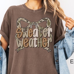 May include: Espresso-colored t-shirt with the words "Sweater Weather" in a decorative font, accented by a camouflage bow. The shirt is part of a clothing collection.
