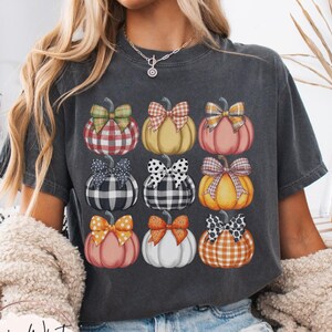 Peut inclure: T-shirt gris fonc&eacute; avec une grille de neuf citrouilles aux motifs et n&oelig;uds vari&eacute;s. Les citrouilles sont orange, rose, jaune et blanc. Le texte "Lovin What U Wear" est visible. Le t-shirt est &eacute;tiquet&eacute; "Pepper".