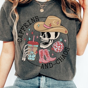 Pu&ograve; includere: Una maglietta grigia con una grafica di uno scheletro che indossa un cappello da cowboy e tiene una tazza di caff&egrave;. Il testo "CAFFEINE AND CHAOS" &egrave; scritto intorno allo scheletro.