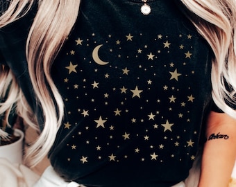 May include: A black t-shirt with a gold crescent moon and stars design. The shirt is worn by a person with long blonde hair and white jeans.