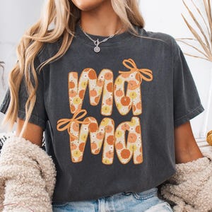 May include: A dark gray t-shirt with the word "Mama" in orange letters filled with a pumpkin pattern. The shirt has orange bows above the first "Ma" and below the second "Ma". The shirt is part of the Lovin What U Wear Etsy shop.