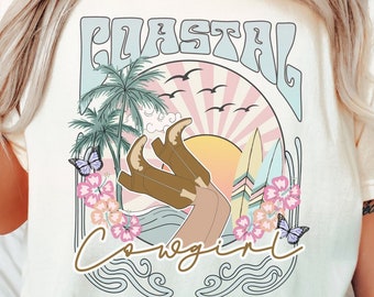 以下が含まれることがあります： レトロなグラフィックデザインのクリーム色のTシャツ。デザインは、ヤシの木、夕日、蝶とともに、様式化されたフォントで「Coastal Cowgirl」という言葉を特徴としています。シャツはリラックスしたフィット感です。
