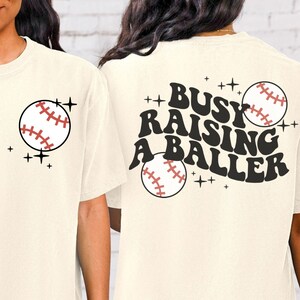 May include: A cream coloured t-shirt with a baseball graphic on the left side and the text "Busy Raising A Baller" with a baseball graphic on the back.