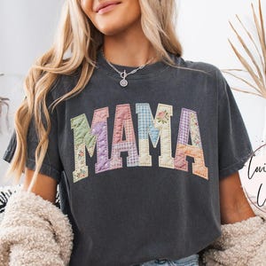 May include: A dark grey t-shirt featuring the word "MAMA" in patchwork lettering. The letters are crafted from diverse floral and patterned fabrics. The shirt has a relaxed fit and short sleeves. The text "Lovin What U Wear" is visible on the right side of the shirt.
