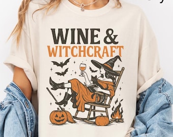 May include: Ivory-colored t-shirt with the words "WINE & WITCHCRAFT" in brown and orange. The graphic features a witch in a rocking chair, holding a wine glass, with pumpkins, bats, and a bonfire.