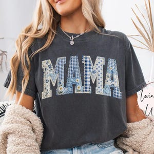 May include: Dark gray t-shirt with the word "MAMA" in patchwork letters. The letters are a mix of floral and gingham patterns in blue and white. The shirt is part of the "Lovin What U Wear" shop collection.