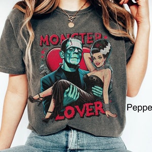 May include: A gray t-shirt with a graphic print of Frankenstein and his bride. The graphic includes the text "Monster Lover" and a red heart. The model is wearing blue jeans and white sneakers.