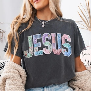 Può includere: T-shirt grigio scuro con la scritta "JESUS" in un design patchwork. Le lettere sono in vari colori pastello e motivi floreali. La maglietta è indossata con pantaloncini di jeans e un cardigan color crema.