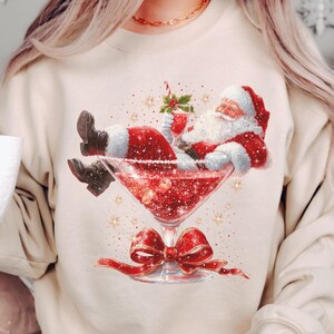 May include: Sand-colored sweatshirt featuring a graphic of Santa Claus relaxing in a cocktail glass. The design includes a red bow, glitter, and the text "Lovin What U Wear". A festive and fun holiday-themed apparel item.