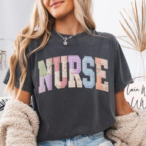 Puede incluir: Camiseta gris oscuro con la palabra "NURSE" en un diseño de patchwork en el pecho. Las letras son de varios colores pastel. La modelo lleva pantalones cortos vaqueros y una rebeca color crema.