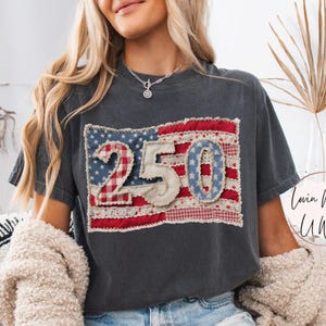 May include: Dark gray t-shirt with a patriotic design. The shirt features a patchwork flag with the numbers "250" in the center. The text "Lovin What U Wear" is visible on the right side of the shirt. The word "Pepper" is also visible.
