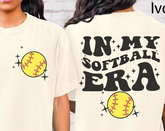 May include: Ivory t-shirt with short sleeves and a graphic print of a softball with the text "IN MY SOFTBALL ERA" on the back.