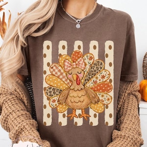May include: A brown t-shirt featuring a graphic of a turkey with a plaid bow on its head. The turkey is centred over a background of vertical stripes with polka dots. The turkey's feathers are decorated with various patterns including floral, leopard print, and plaid.