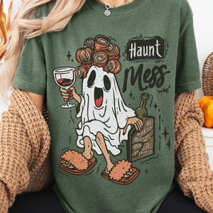 May include: Moss green t-shirt with a cartoon ghost design. The ghost is holding a wine glass and wearing curlers and slippers. The text "Haunt Mess" is above the ghost. A bottle and a sign are also in the design.