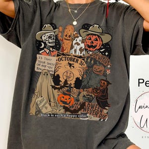 May include: Pepper-colored graphic tee featuring a Halloween-themed collage. The design includes a skeleton, a pumpkin, a ghost, and the text "October 31". The shirt also has the text "Black is such a happy color".