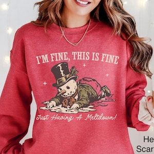 以下が含まれることがあります： 「I'M FINE, THIS IS FINE」の文字と雪だるまのグラフィックがプリントされた、ヘザー・スカーレット・レッドのスウェットシャツ。雪だるまはシルクハットをかぶり、横たわっています。「Just Having A Meltdown!」の文字が下にあります。