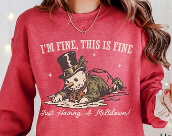 Può includere: Felpa Heather Scarlet Red con la scritta "I'M FINE, THIS IS FINE" e una grafica di pupazzo di neve. Il pupazzo di neve indossa un cappello a cilindro ed è sdraiato. Il testo "Just Having A Meltdown!" è sotto.