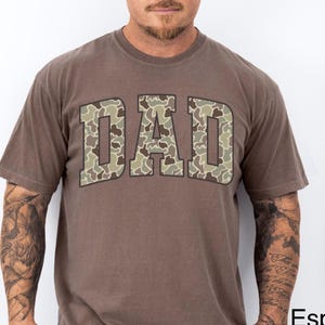 May include: Espresso-colored t-shirt with the word "DAD" in a camouflage pattern across the chest. The shirt has short sleeves and a crew neck. The model is wearing blue jeans. The shirt is part of the Lovin What U Wear Etsy shop.