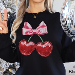 May include: Black sweatshirt with a pink bow and two red cherries with sequins. The cherries have green stems. The text "Lovin' What U Wear" is visible in the bottom right corner.
