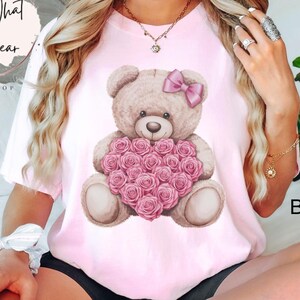 May include: A pink t-shirt with a graphic of a brown teddy bear holding a heart-shaped bouquet of pink roses. The teddy bear is wearing a pink bow. The text "Blossom" is visible in the bottom right corner of the image.
