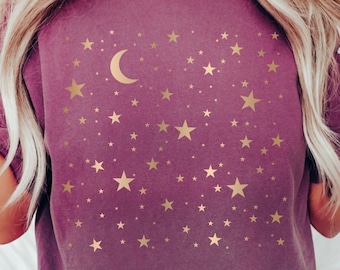 May include: A plum-colored t-shirt featuring a gold crescent moon and numerous gold stars of varying sizes. The celestial design is printed on a soft, comfortable fabric. This shirt is a great addition to any wardrobe.